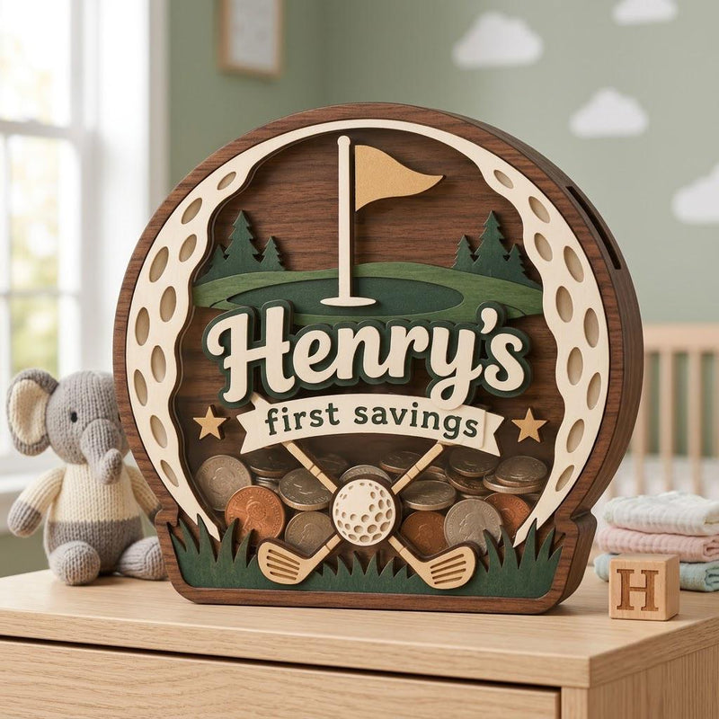 May include: A circular wooden piggy bank shaped like a golf ball with a dark wood frame. The center features a green golf course, a flag, and two crossed golf clubs. It displays the text Henry's first savings in white lettering.