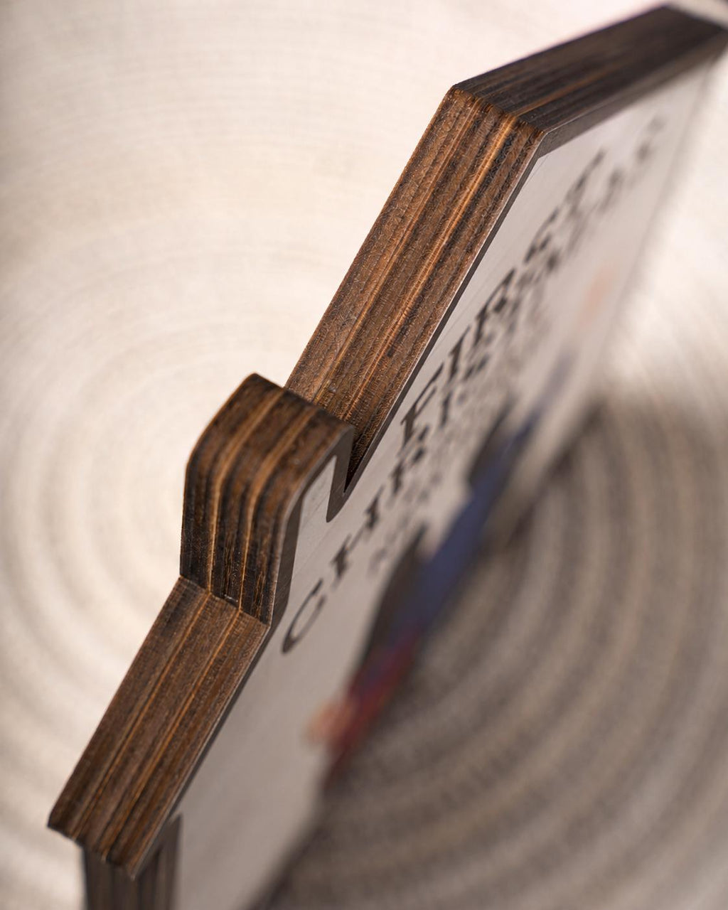 May include: A wooden sign with the words "FIRST CHRISTMAS" visible. The sign has a dark brown, layered edge and a notch on the side. The background is a light, textured surface.