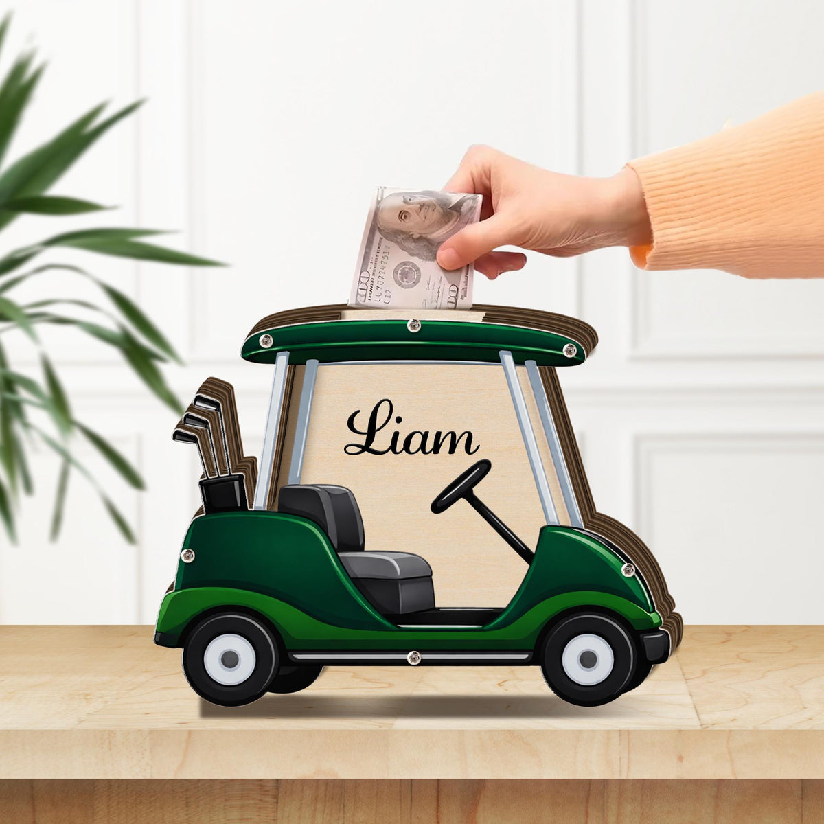 May include: A green and brown wooden golf cart-shaped bank with the name "Liam" on the side. A hand is inserting a $100 bill into the slot on top. The bank has a steering wheel, seat, and golf club details.
