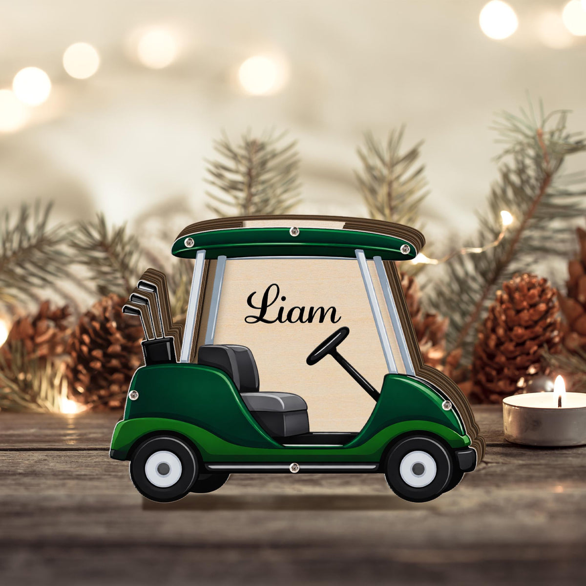 May include: A wooden golf cart ornament in green and brown, with the name "Liam" scripted on the center. The cart features a steering wheel, seats, and golf clubs. The background includes pine needles and a candle.