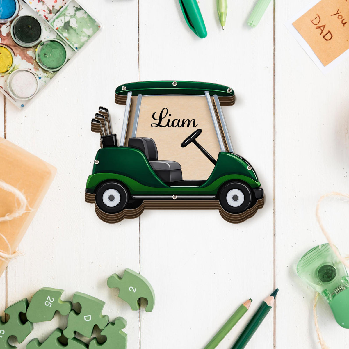 May include: A wooden golf cart-shaped decoration in shades of green and brown, with the name "Liam" displayed on the cart. The cart features a steering wheel, seats, and golf clubs. The background includes art supplies and a small card that says "you DAD".