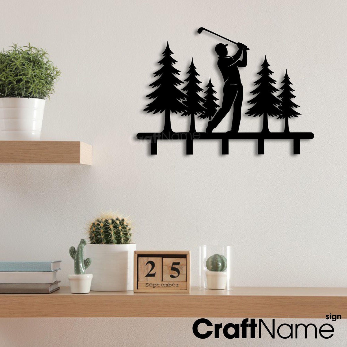 May include: A black metal wall-mounted rack featuring a silhouette of a golfer swinging a club amidst a forest of pine trees. The design includes five downward-facing hooks at the base for hanging items, with a flat, laser-cut metal construction.
