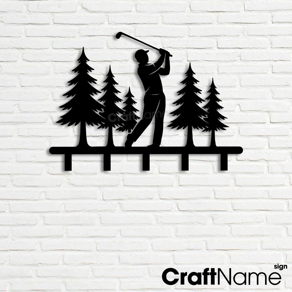 May include: A black metal wall-mounted hook rack featuring a silhouette of a golfer swinging a club amidst a forest of five pine trees. The design includes a horizontal base bar with five downward-facing hooks for hanging items.