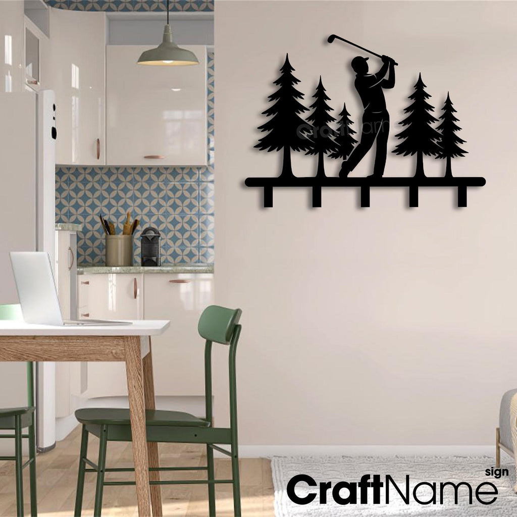 May include: A black metal wall-mounted rack featuring a silhouette of a golfer swinging a club amidst a forest of pine trees. The design includes five downward-facing hooks along the bottom edge for hanging items, presented as a flat decorative piece.