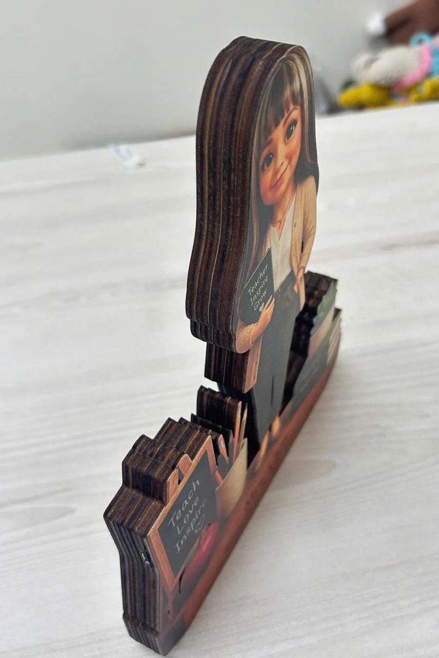 May include: A layered wooden cutout decoration featuring a cartoon-style teacher holding a book with the text "Teach Inspire Grow" and a small chalkboard sign with the same text. The piece has visible dark wood grain edges and a multi-layered design.