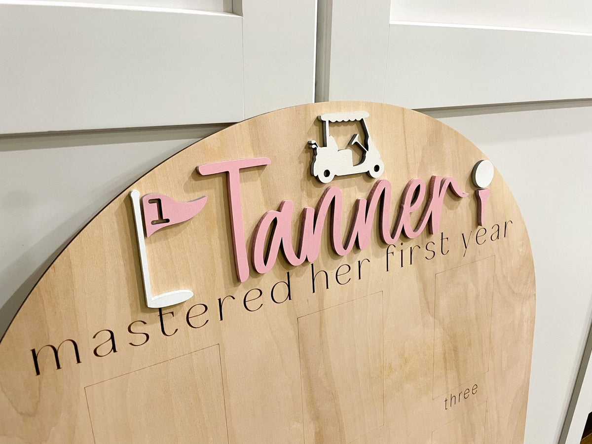 May include: A light wood sign with a pink and white golf theme. The sign says "Tanner mastered her first year" with a golf cart, golf flag, and golf ball.