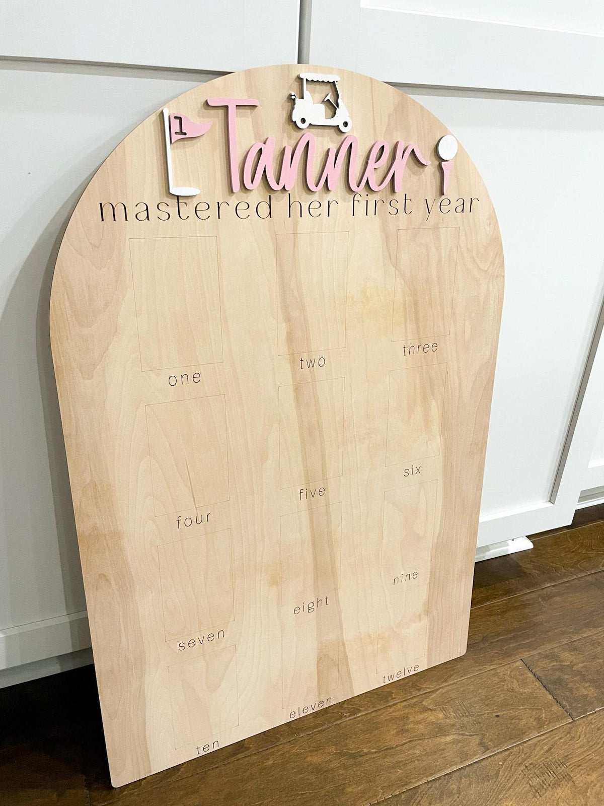 May include: A wooden sign with a pink golf cart and flag, the name Tanner, and the text "mastered her first year". The sign has 12 blank squares with the numbers one through twelve written below each square.