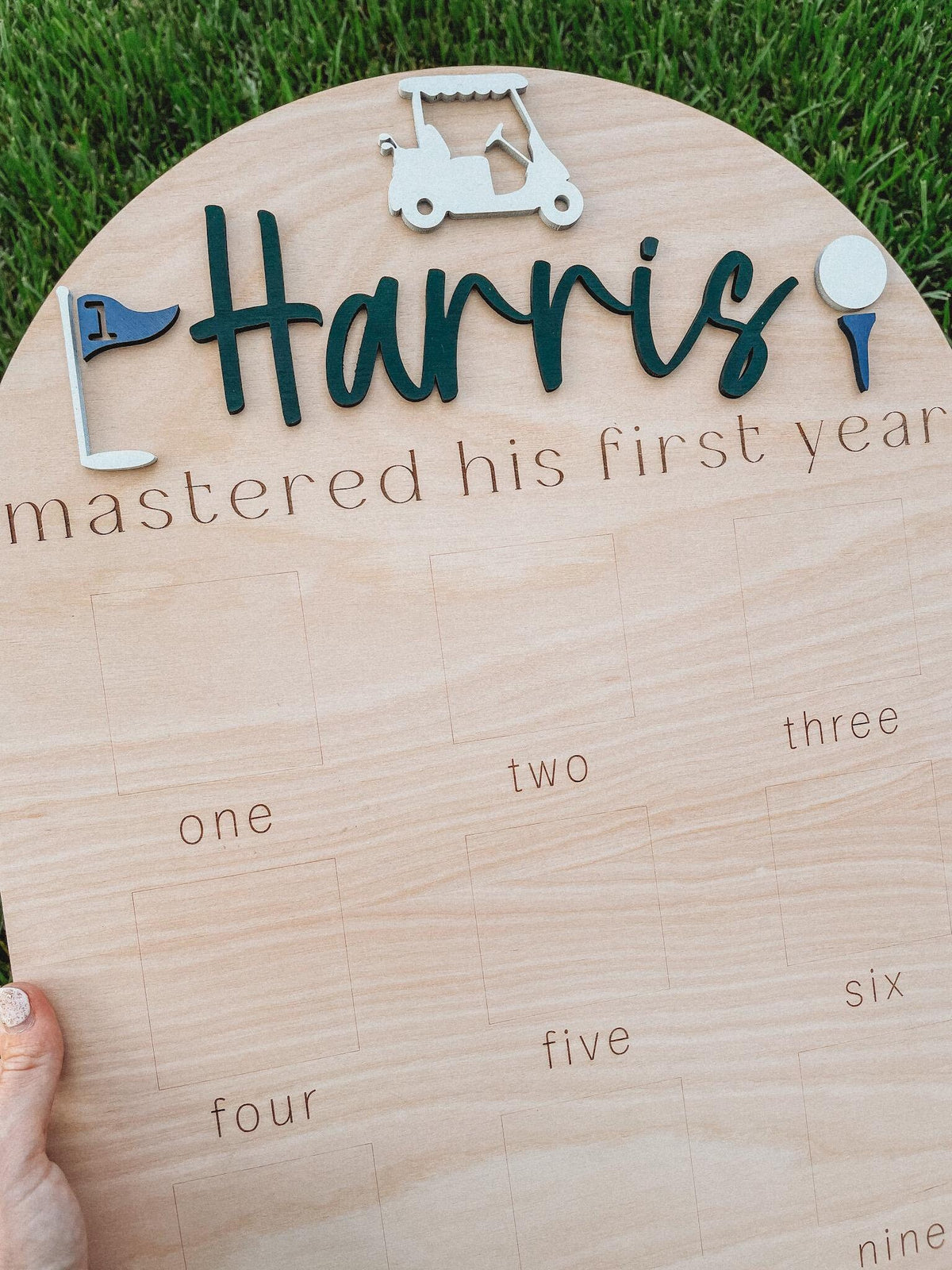May include: A wooden golf-themed growth chart with the name "Harris" and the text "mastered his first year". The chart has nine squares with the numbers one through nine written below each square.