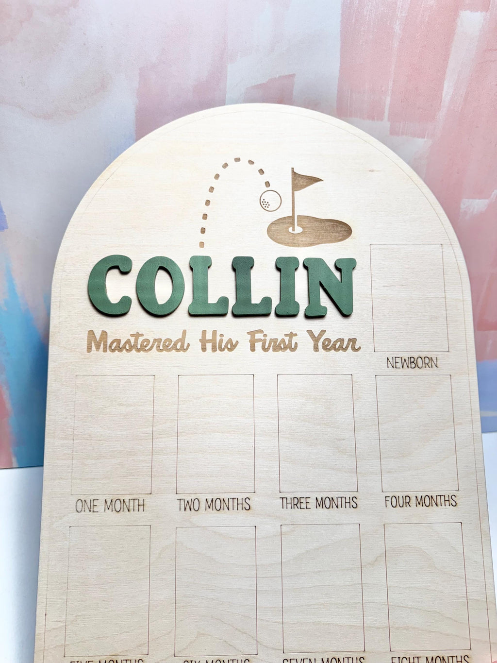 May include: Wooden milestone board with the name "COLLIN" in green letters. The board features a golf-themed design and spaces to document a baby's first year, including "Newborn" and "One Month" to "Eight Months".
