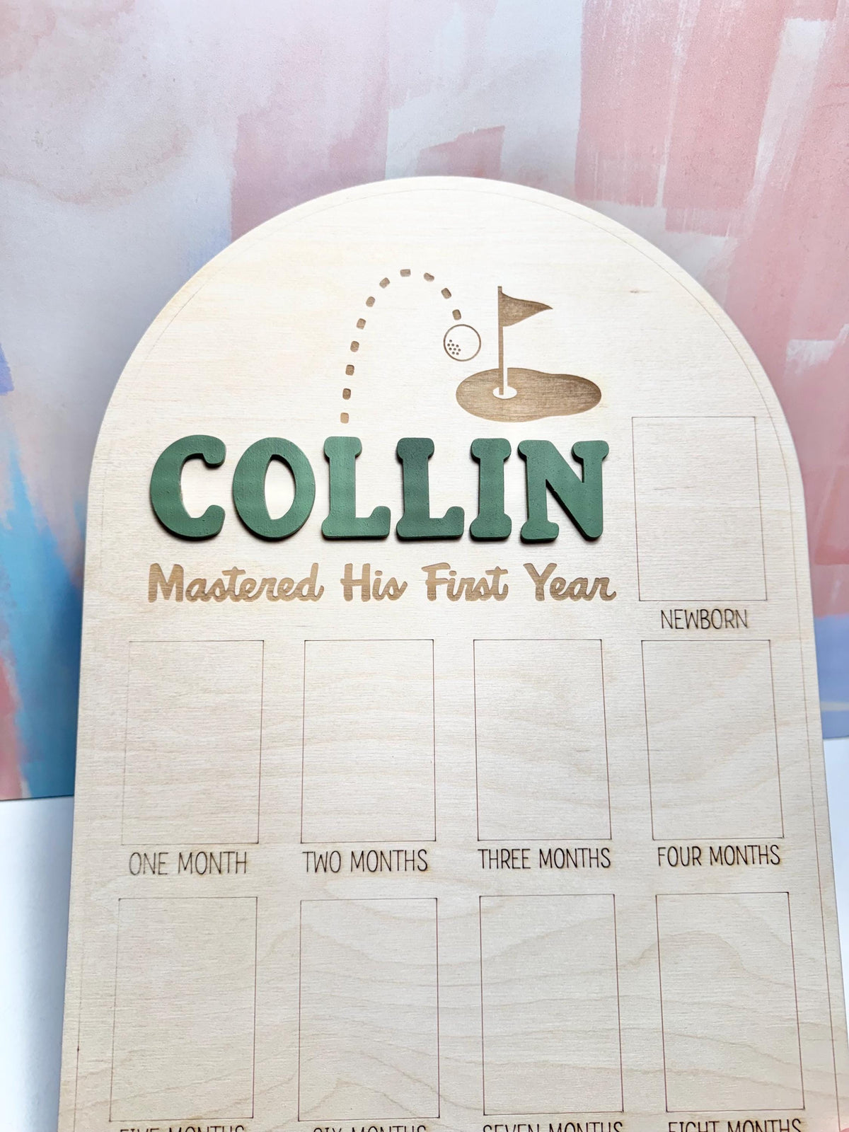 May include: Wooden milestone board with the name "COLLIN" in green letters. The board features a golf-themed design and spaces to document a baby's first year, including "Newborn" and "One Month" to "Eight Months".