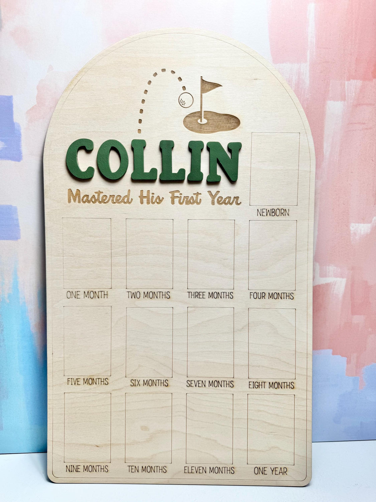 May include: Wooden milestone board with the name "COLLIN" in green letters, and the phrase "Mastered His First Year". The board has a golf-themed design and spaces for monthly photos, from newborn to one year.