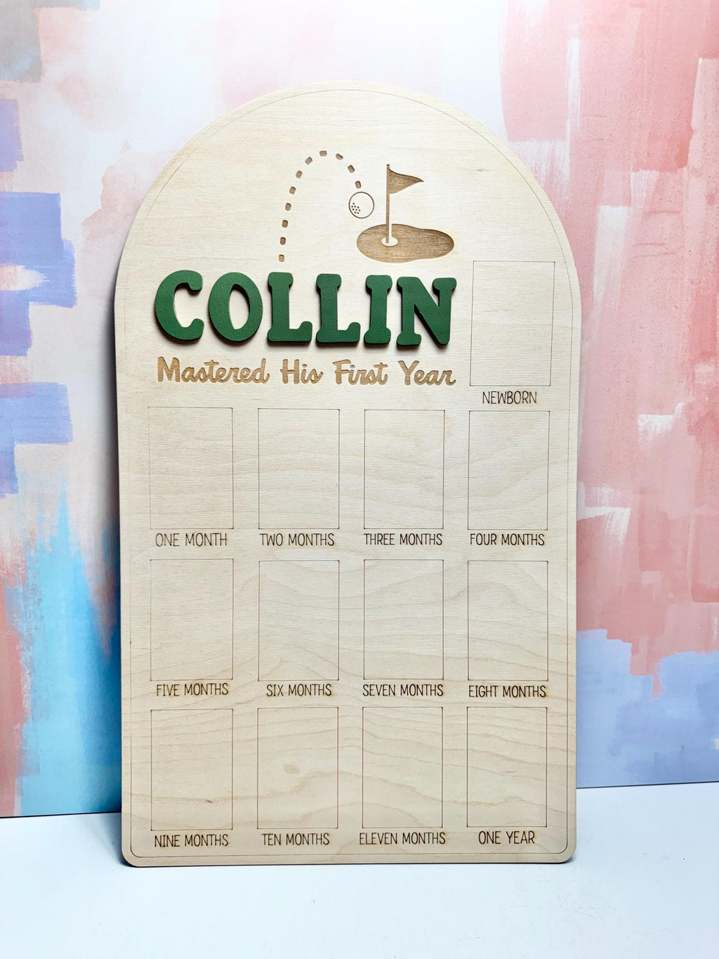 May include: A wooden milestone board with the name "COLLIN" in green letters. The board features a golf-themed design with a golf ball and flag, and spaces for monthly photos from newborn to one year. The text "Mastered His First Year" is also included.