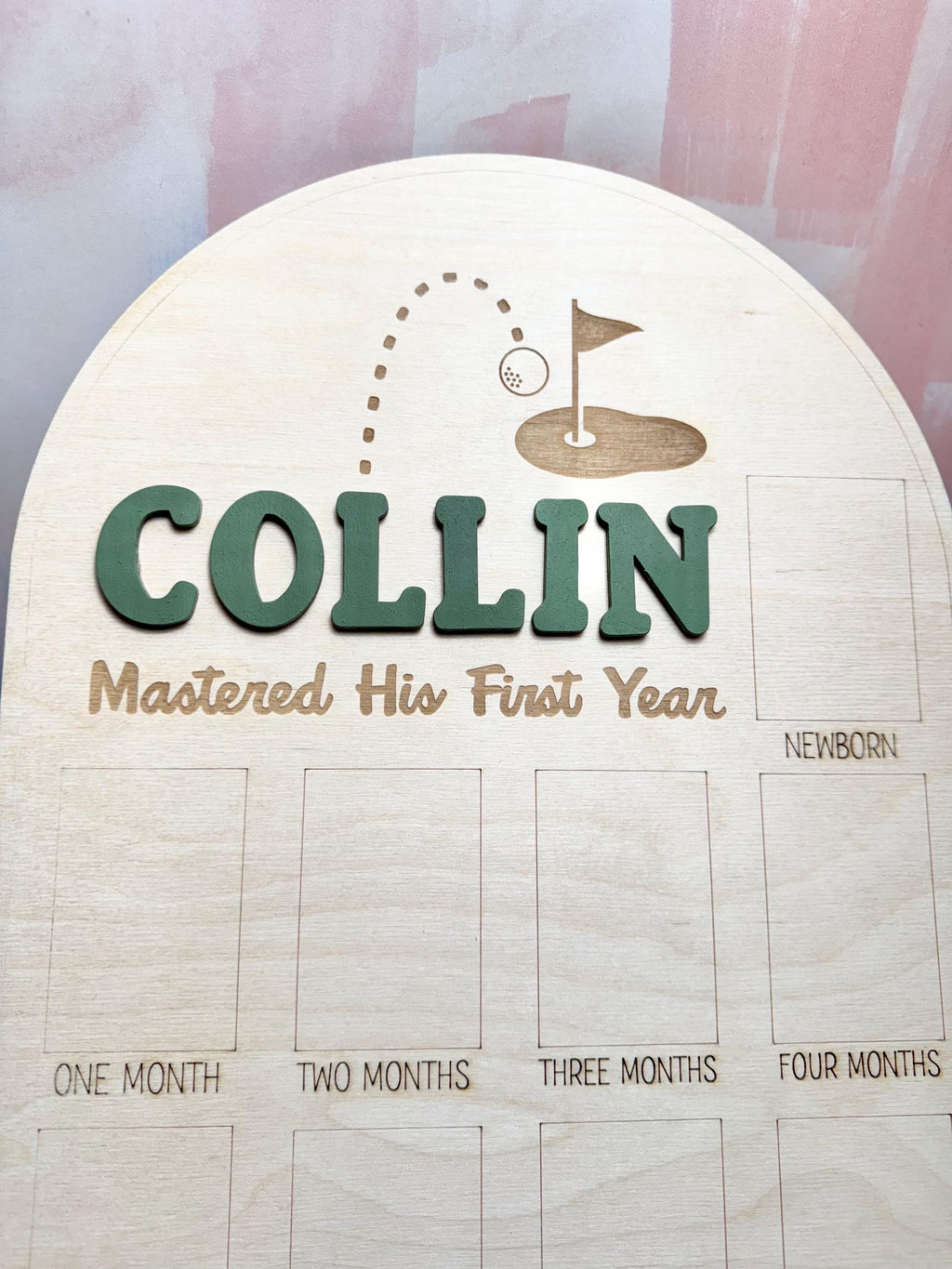 May include: Wooden milestone board with the name "Collin" in green letters. The board features a golf-themed design with a golf ball and flag. It includes spaces for monthly photos and the text "Mastered His First Year."