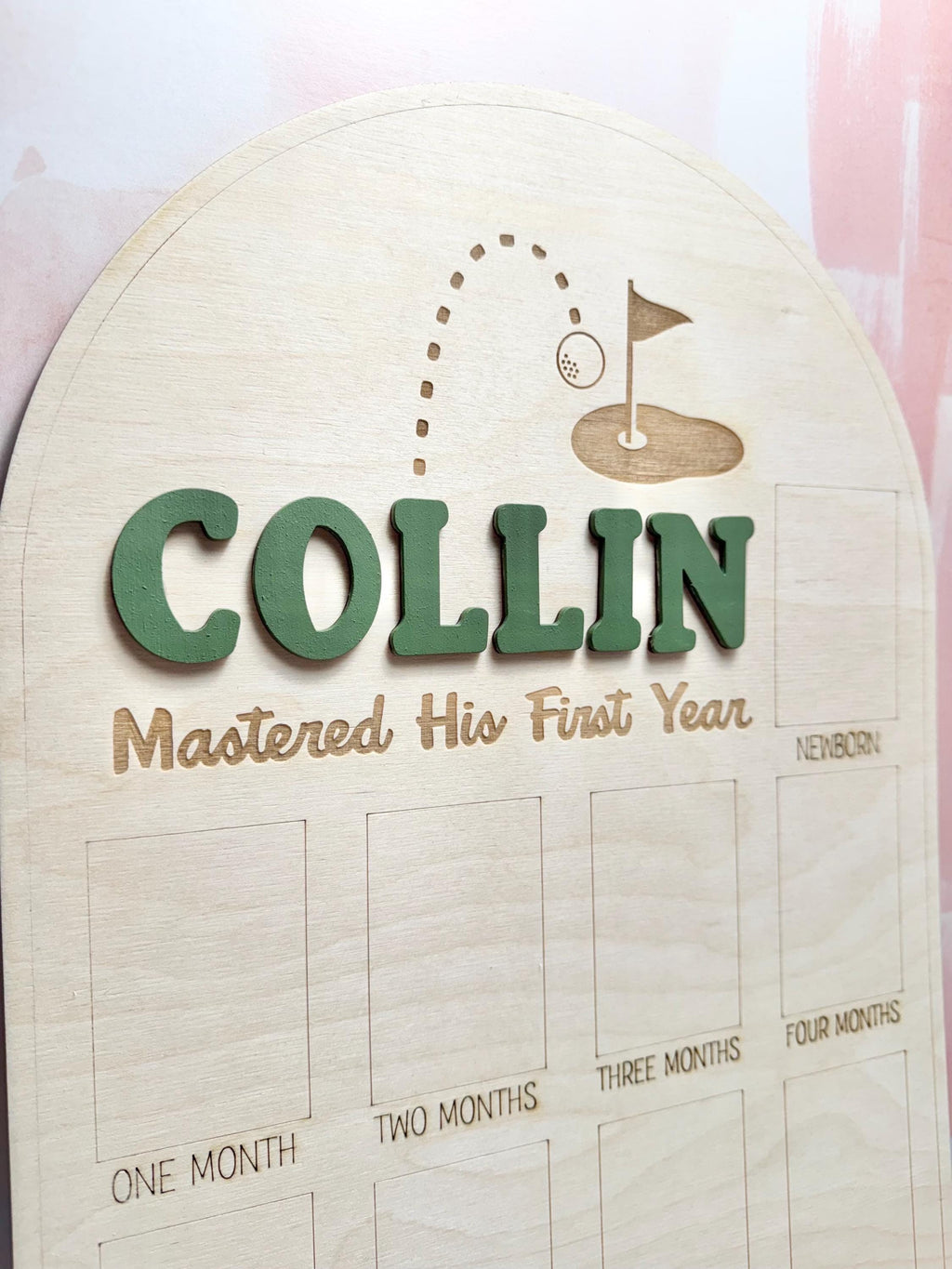 May include: Wooden milestone board with the name "COLLIN" in green 3D letters. The board features a golf-themed design with a golf ball and flag, and the phrase "Mastered His First Year". It includes spaces for monthly photos.