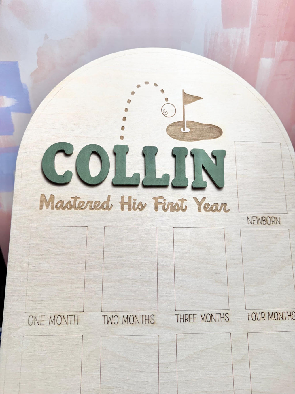 May include: Wooden milestone board with the name "COLLIN" in green 3D letters. The board features a golf-themed design with a ball and flag, and the text "Mastered His First Year." It includes spaces for monthly photos.