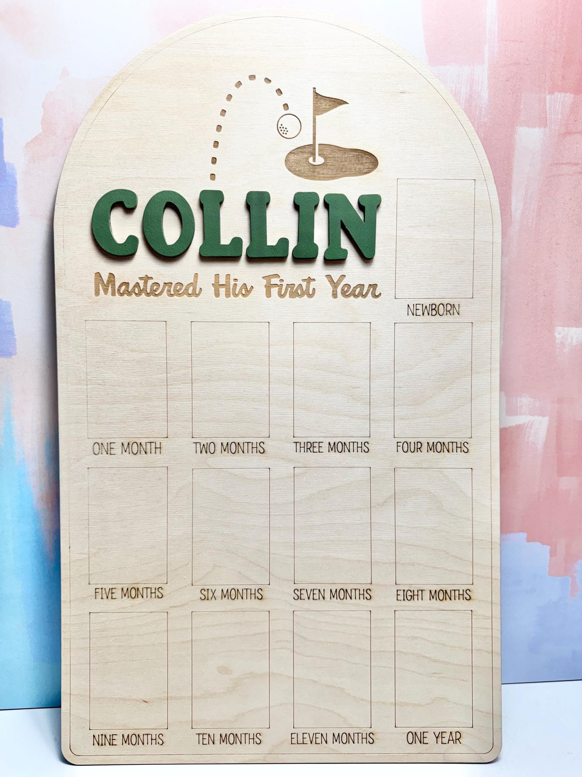 May include: Wooden milestone board with the name "COLLIN" in green, and the phrase "Mastered His First Year." Features a golf-themed design at the top. Includes spaces for monthly photos from newborn to one year.