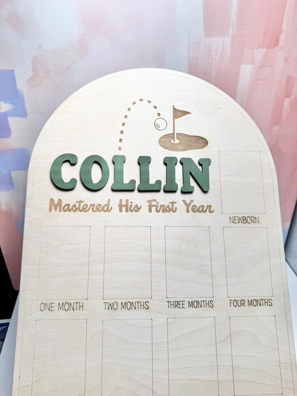May include: Wooden milestone board with the name "Collin" in green, and the phrase "Mastered His First Year". The top features a golf-themed design. Includes spaces for photos labeled "Newborn" through "Twelve Months".