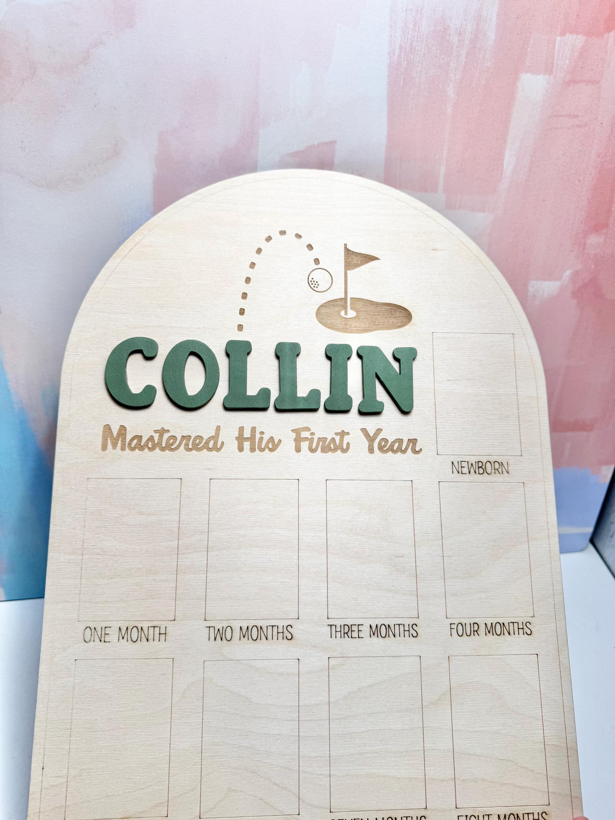 May include: Wooden milestone board with the name "COLLIN" in green, and the phrase "Mastered His First Year". Features a golf-themed design and spaces for monthly photos. The board is made of light-colored wood.