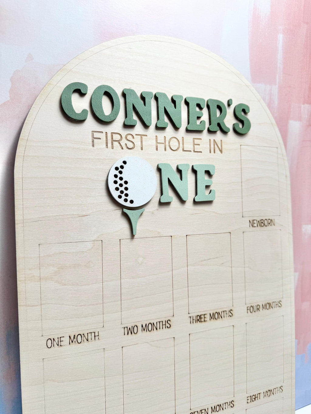 May include: Wooden milestone board with the text "CONNER'S FIRST HOLE IN ONE" in green. A golf ball graphic is featured. The board has spaces for photos labeled "ONE MONTH" to "EIGHT MONTHS" and "NEWBORN".