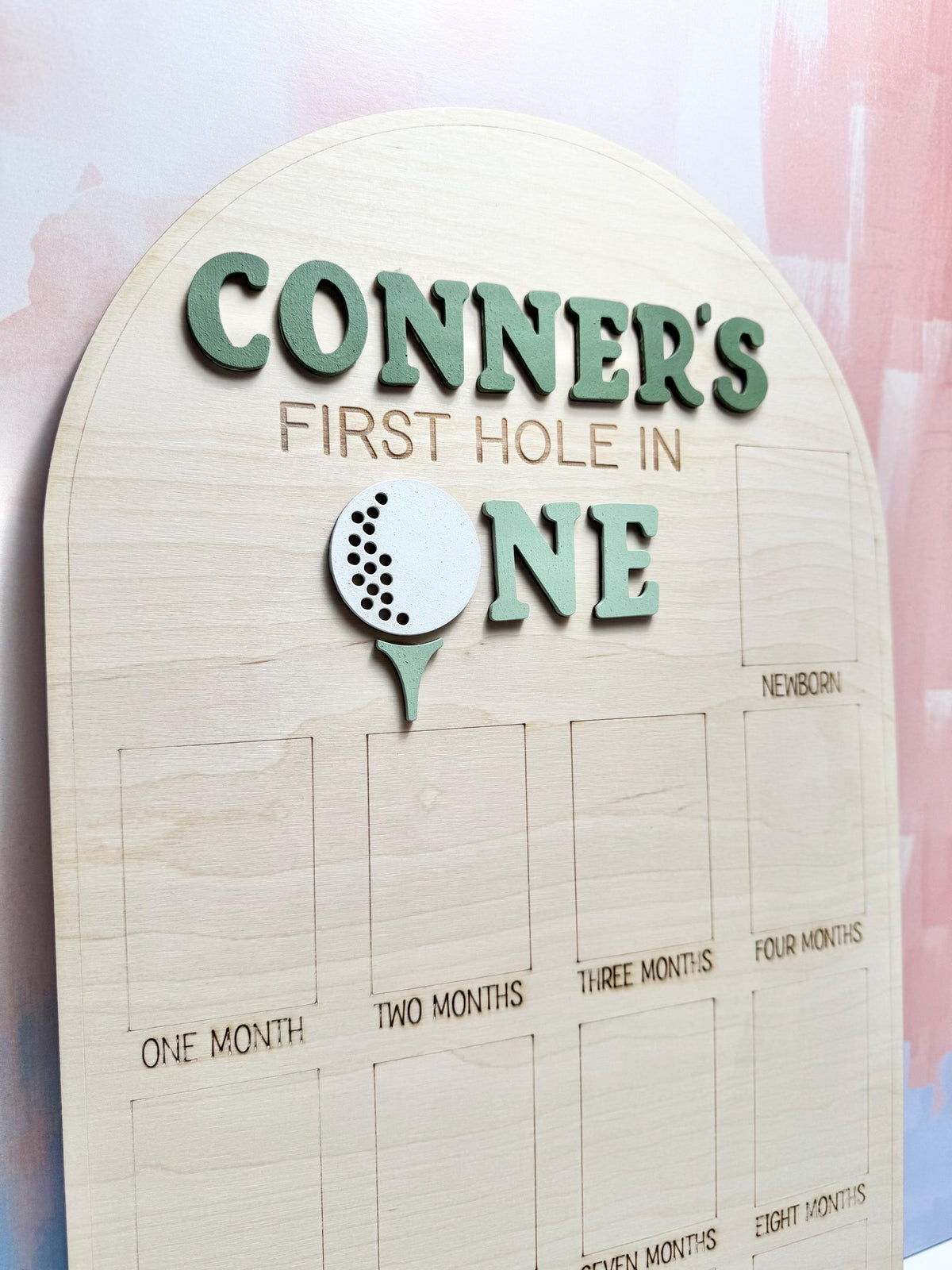 May include: Wooden milestone board with the text "CONNER'S FIRST HOLE IN ONE" in green. A golf ball graphic is featured. The board has spaces for photos labeled "ONE MONTH" to "EIGHT MONTHS" and "NEWBORN".