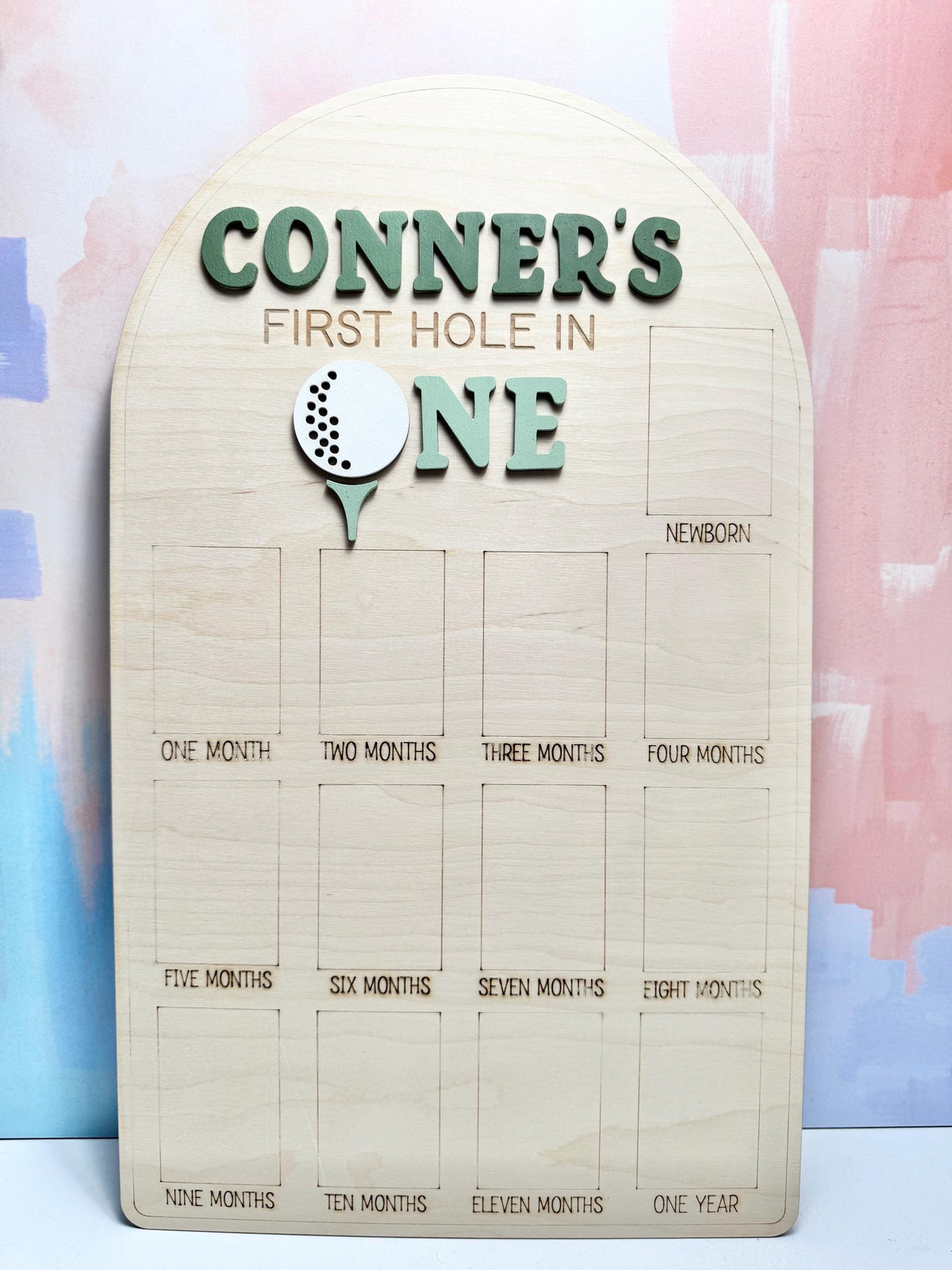 May include: A wooden milestone board with the text "CONNER'S FIRST HOLE IN ONE" in green. It features a golf ball graphic and spaces for monthly photos from newborn to one year. The board is made of light-colored wood.