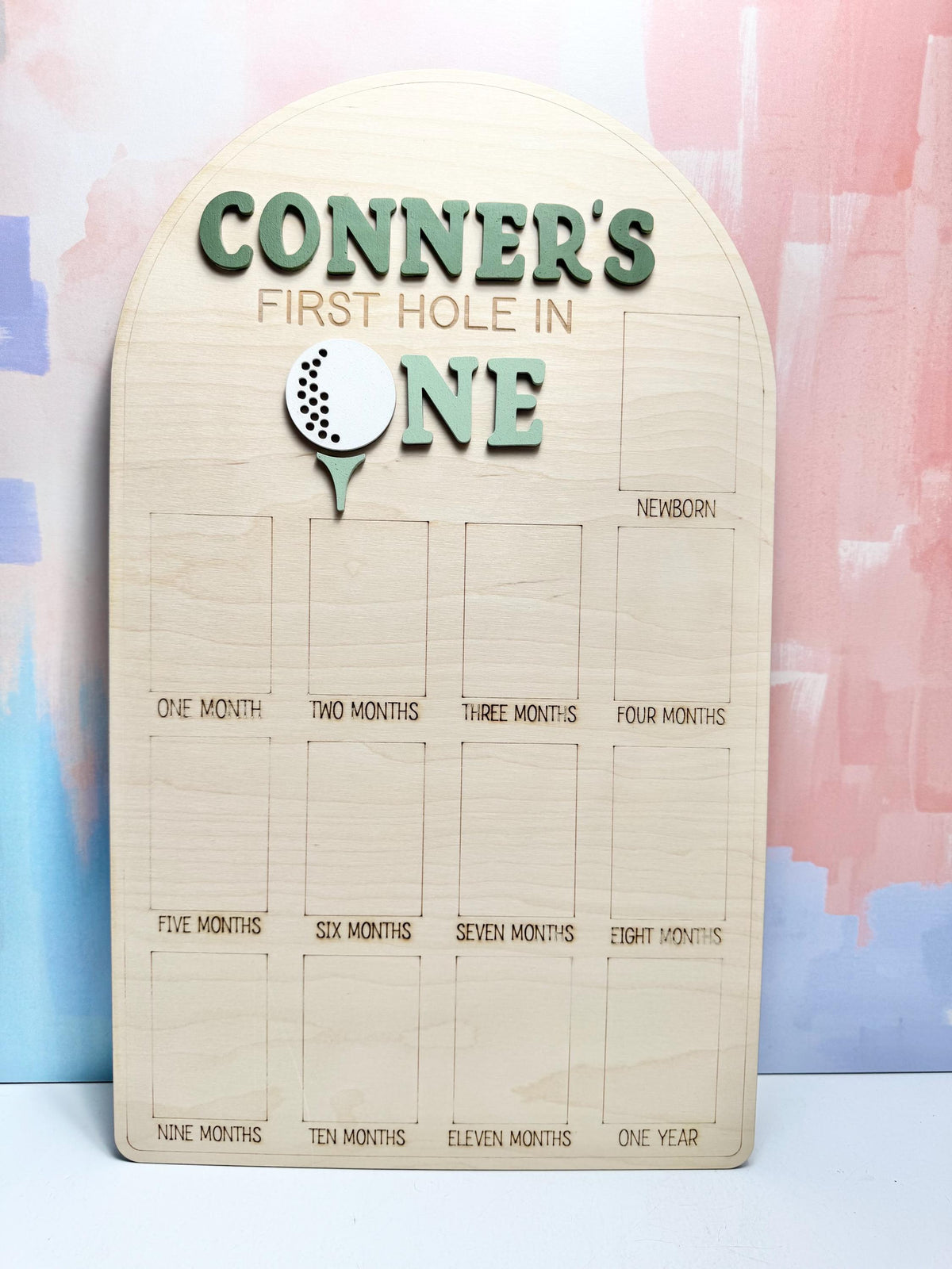 May include: Wooden milestone board with the text "CONNER'S FIRST HOLE IN ONE." Features a golf ball graphic. Includes spaces for monthly milestones from newborn to one year. The board is made of light-colored wood.