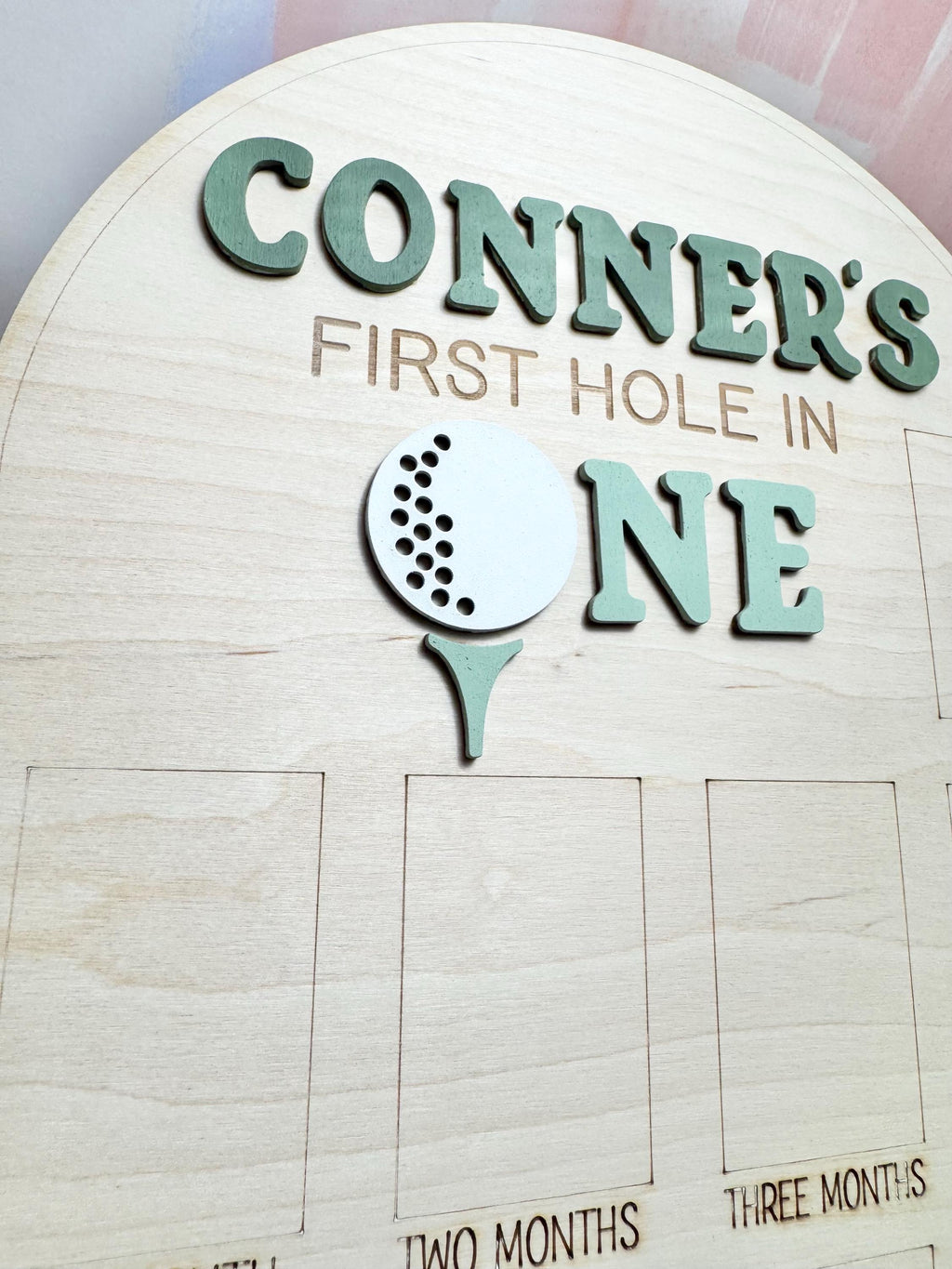 May include: Wooden sign with the text "CONNER'S FIRST HOLE IN ONE" in green letters. A white golf ball and green tee are also featured. The sign has spaces for monthly milestones.