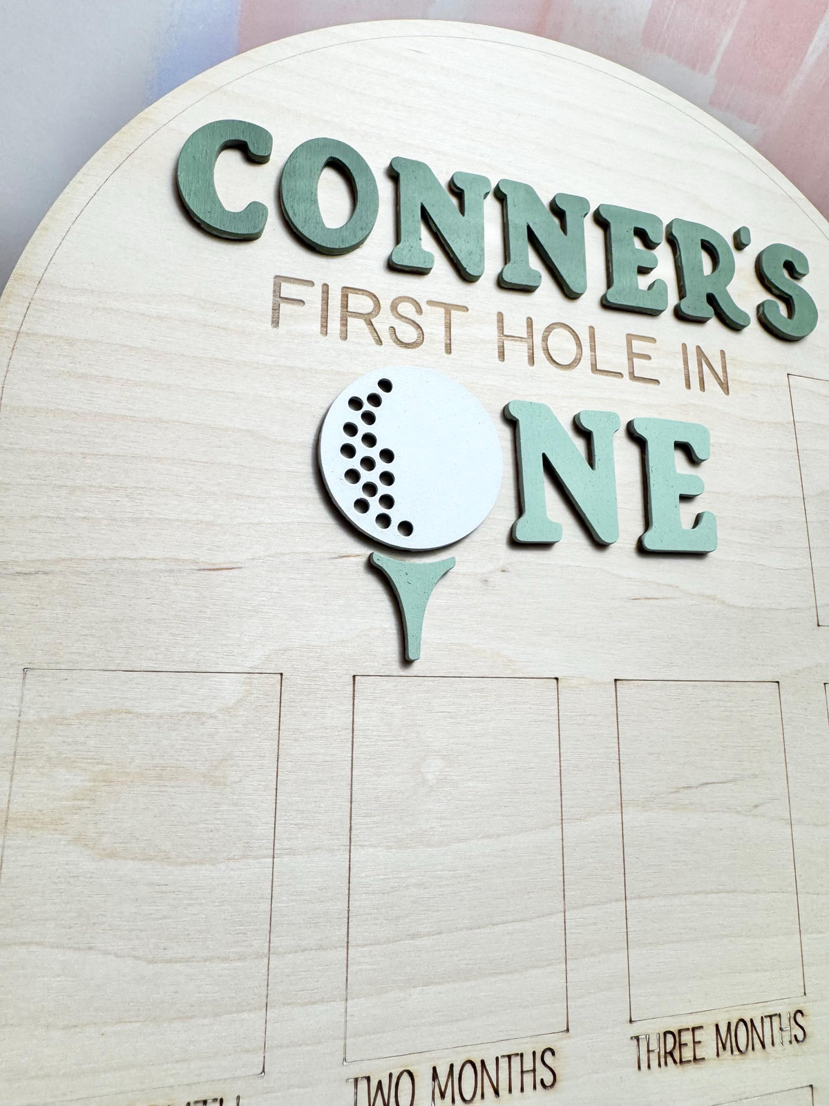 May include: Wooden sign with the text "CONNER'S FIRST HOLE IN ONE" in green letters. A white golf ball and green tee are also featured. The sign has spaces for monthly milestones.