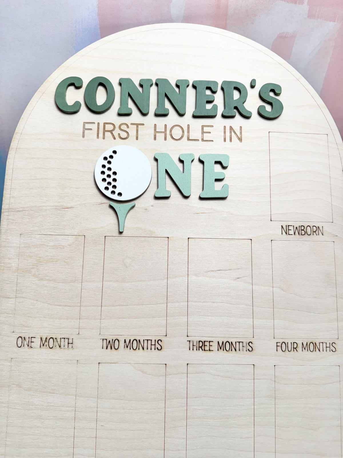 May include: Wooden milestone board with the text "CONNER'S FIRST HOLE IN ONE" in green letters. A golf ball and tee graphic are below the text. The board has spaces for photos labeled "NEWBORN", "ONE MONTH" through "FOUR MONTHS".