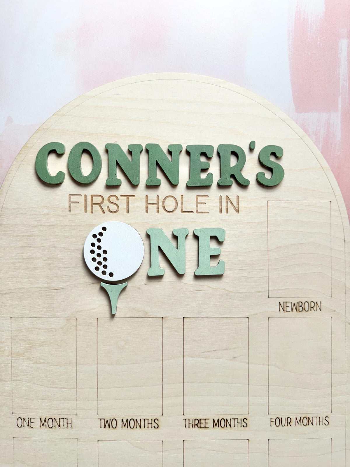 May include: A wooden milestone board with the text "CONNER'S FIRST HOLE IN ONE" in green letters. A golf ball graphic is above the word "ONE". The board has spaces for monthly photos, labeled "ONE MONTH" through "FOUR MONTHS" and "NEWBORN".