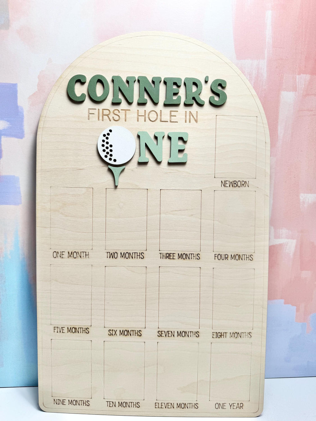 May include: Wooden milestone board with the text "CONNER'S FIRST HOLE IN ONE" in green letters. A golf ball and tee graphic is featured. The board has spaces for monthly milestones from newborn to one year.