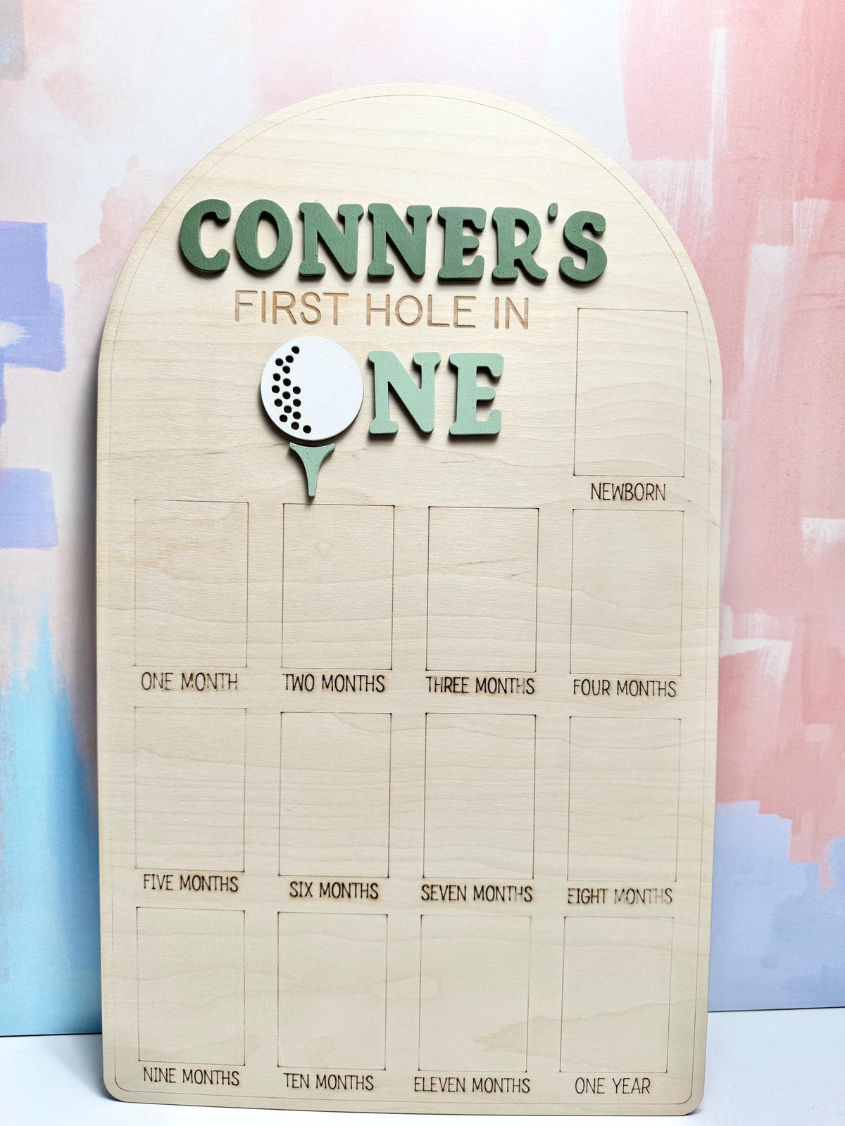 May include: Wooden milestone board with the text "CONNER'S FIRST HOLE IN ONE" in green letters. A golf ball and tee graphic is featured. The board has spaces for monthly milestones from newborn to one year.
