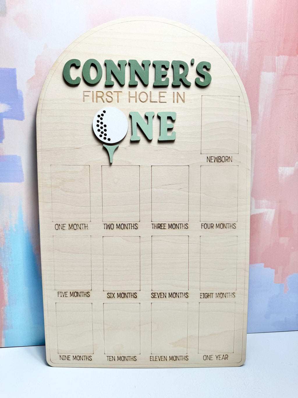 May include: Wooden milestone board with the text "CONNER'S FIRST HOLE IN ONE." Features a golf ball graphic. Includes spaces for monthly photos from newborn to one year. The board is made of light-colored wood.