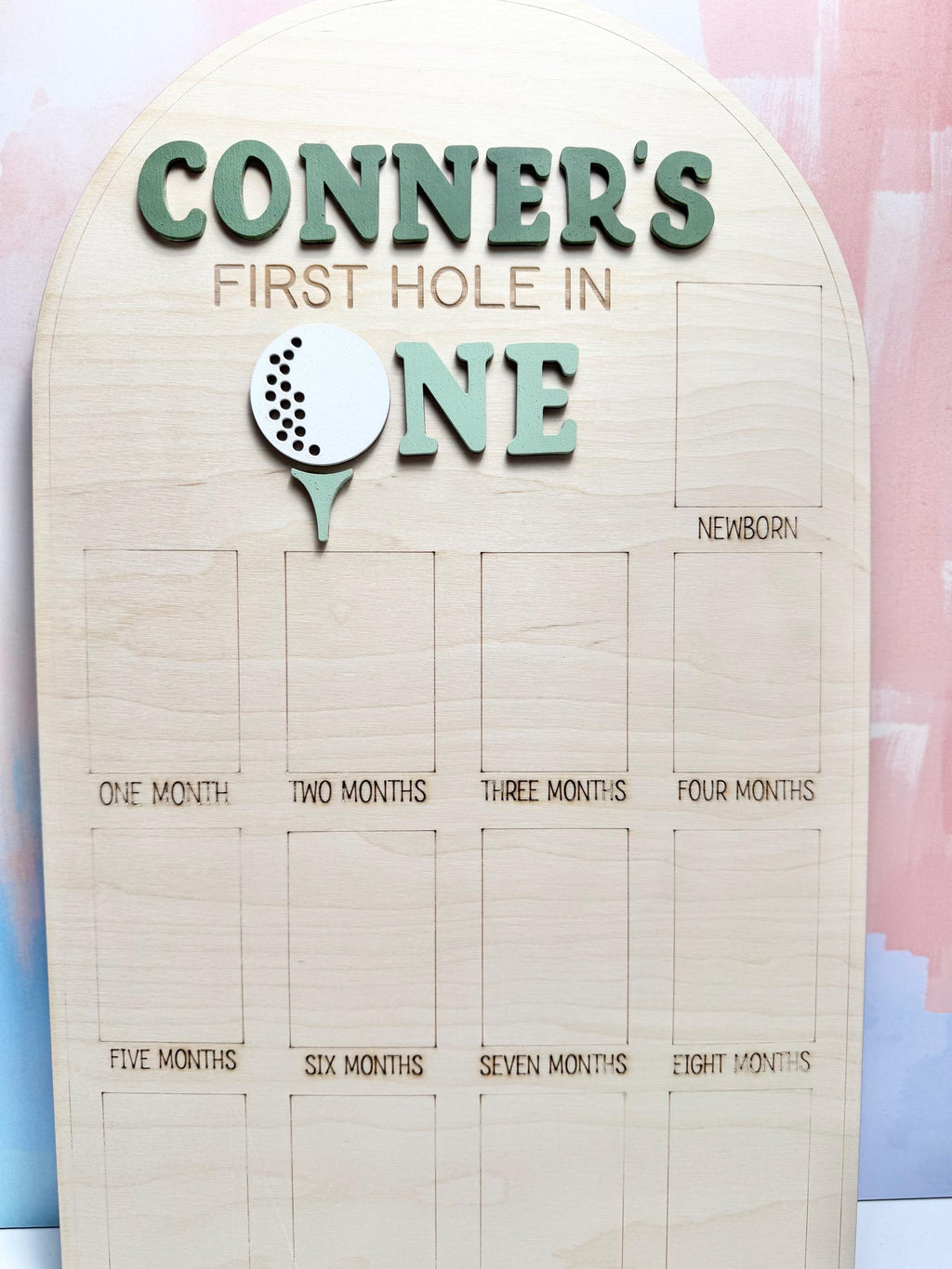 May include: Wooden milestone board with the text "CONNER'S FIRST HOLE IN ONE." The board features a golf ball and tee design. It has spaces for monthly milestones from newborn to eight months. The board is made of light-colored wood.