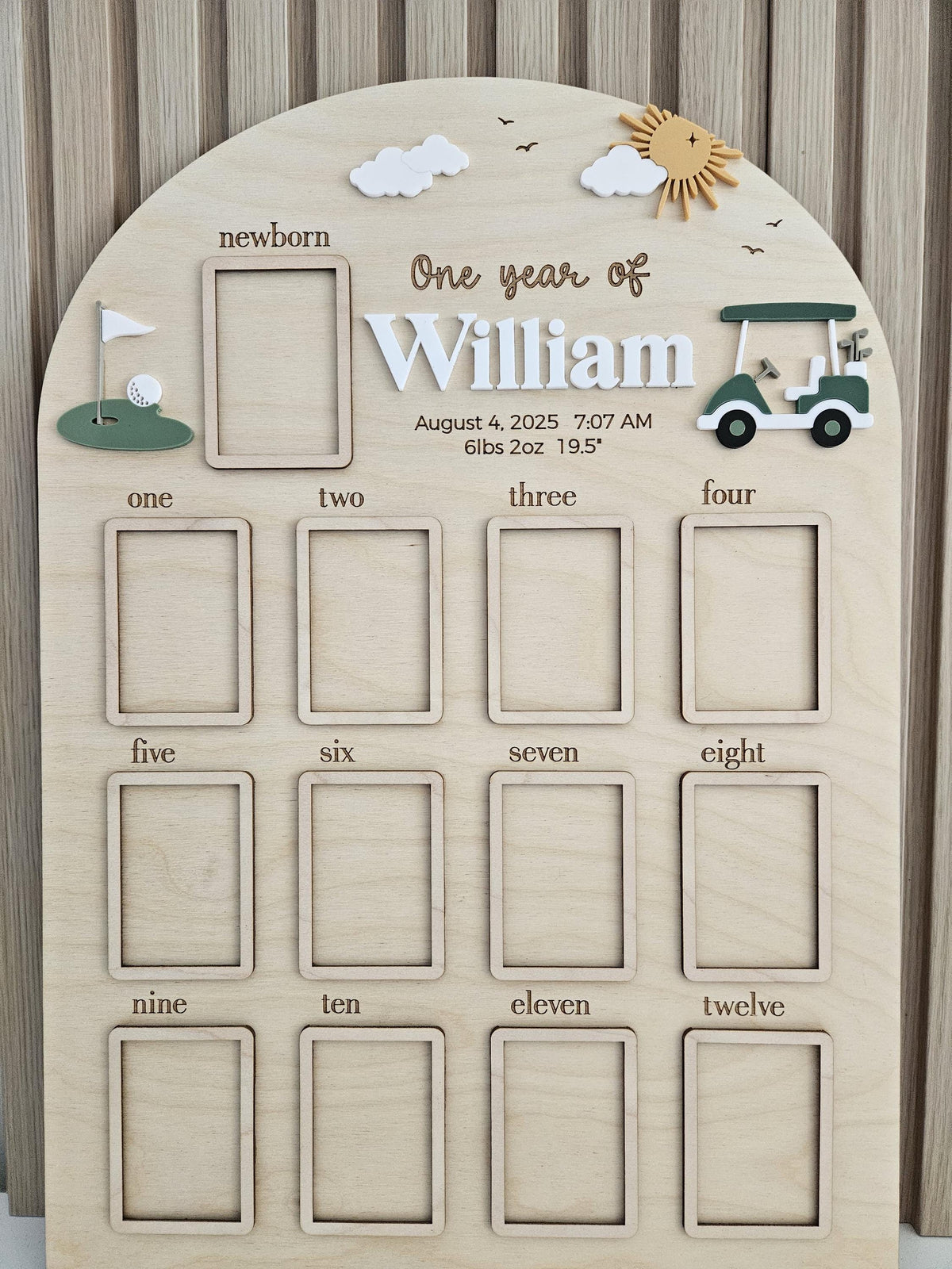 May include: An arched wooden milestone board featuring the text One year of William, August 4, 2025, 7:07 AM, 6lbs 2oz 19.5 inches. It includes a newborn frame, twelve monthly frames, a golf cart, a putting green, clouds, and a sun cutout.