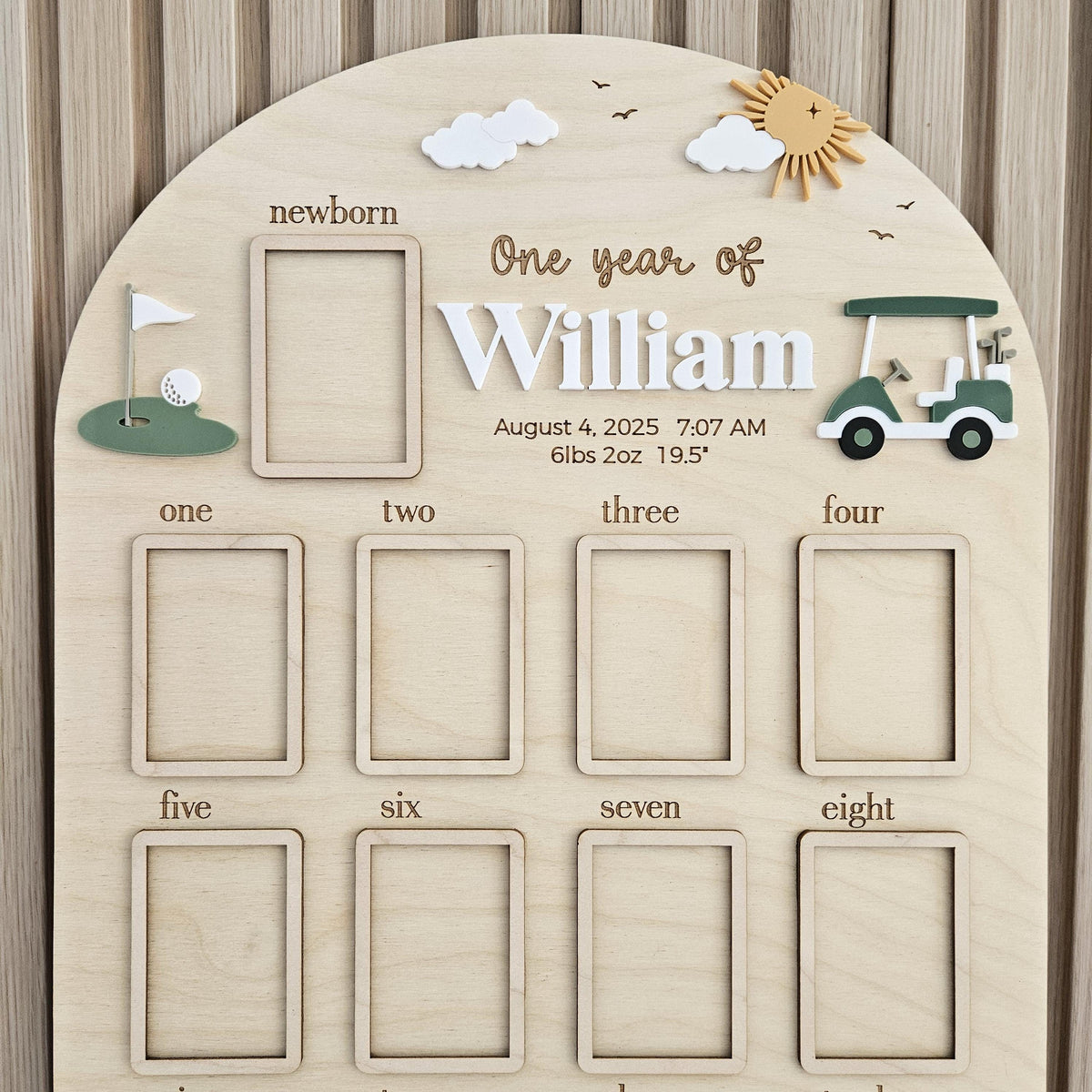 May include: An arched wooden milestone board featuring the text One year of William, August 4, 2025, 7:07 AM, 6lbs 2oz, 19.5 inches. It includes a newborn frame, eight monthly frames, a golf cart, a golf hole with a flag, clouds, and a sun.