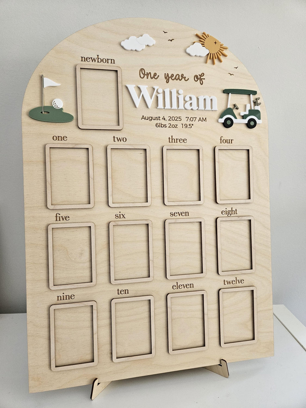May include: An arched wooden milestone board featuring a golf theme with a green putting hole, a golf cart, clouds, and a sun. It includes a frame for a newborn photo, twelve additional frames for monthly photos, and text reading One year of William.