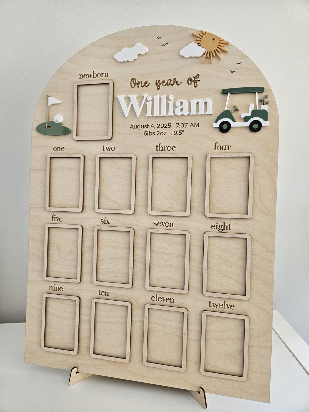 May include: An arched wooden milestone board featuring a golf theme with a green golf cart, a flag on a green, and a sun. It includes thirteen rectangular photo frames labeled from newborn to twelve, with the text One year of William, August 4, 2025, 7:07 AM, 6lbs 2oz 19.5 inches.