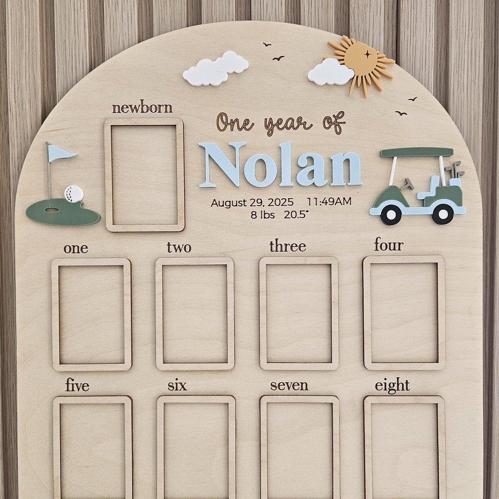May include: An arched wooden milestone board featuring a golf theme with a green golf cart, a putting green, clouds, and a sun. It includes text reading One year of Nolan, August 29, 2025, 11:49AM, 8 lbs, 20.5 inches, and frames for photos from newborn to eight.