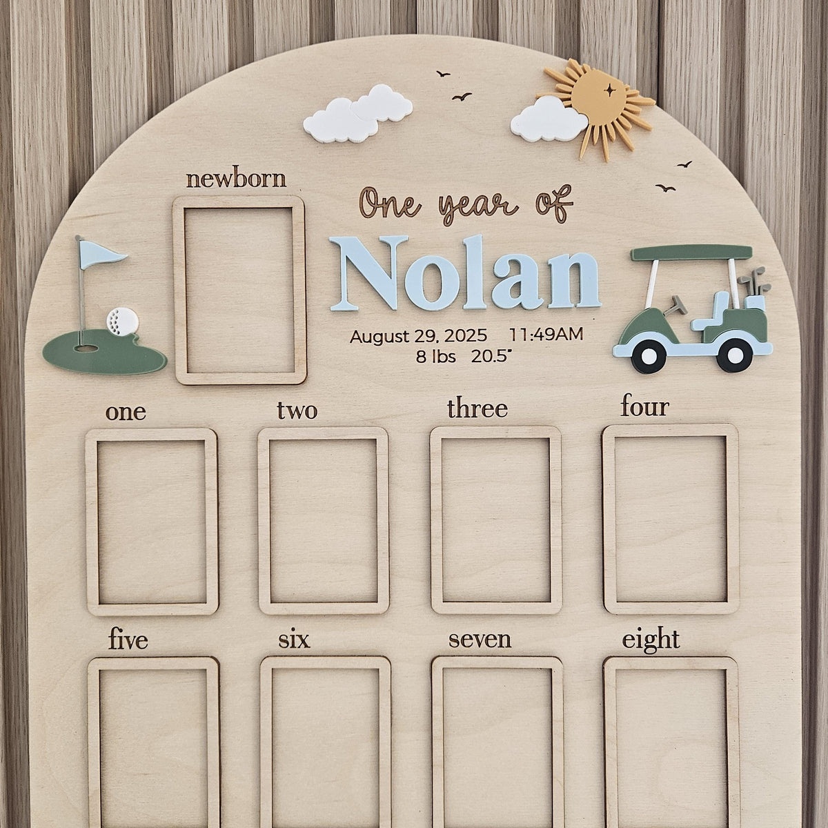 May include: An arched wooden milestone board featuring a golf theme with a green golf cart, a putting green, clouds, and a sun. It includes text reading One year of Nolan, August 29, 2025, 11:49AM, 8 lbs, 20.5 inches, and frames for photos from newborn to eight.