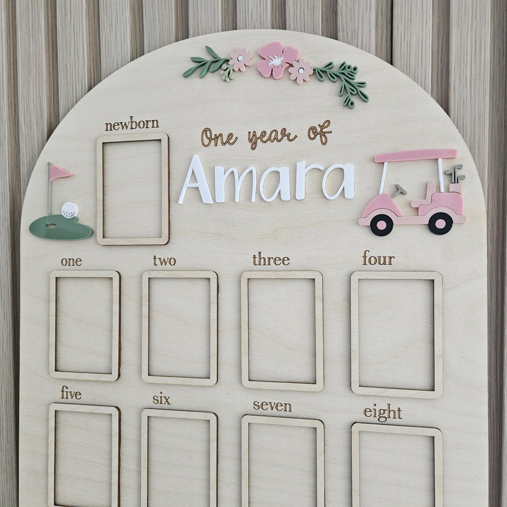 May include: An arched wooden milestone board featuring the text One year of Amara, with a pink golf cart, a green golf hole, and floral accents. It includes twelve rectangular frames labeled from newborn to eight for photo placement.