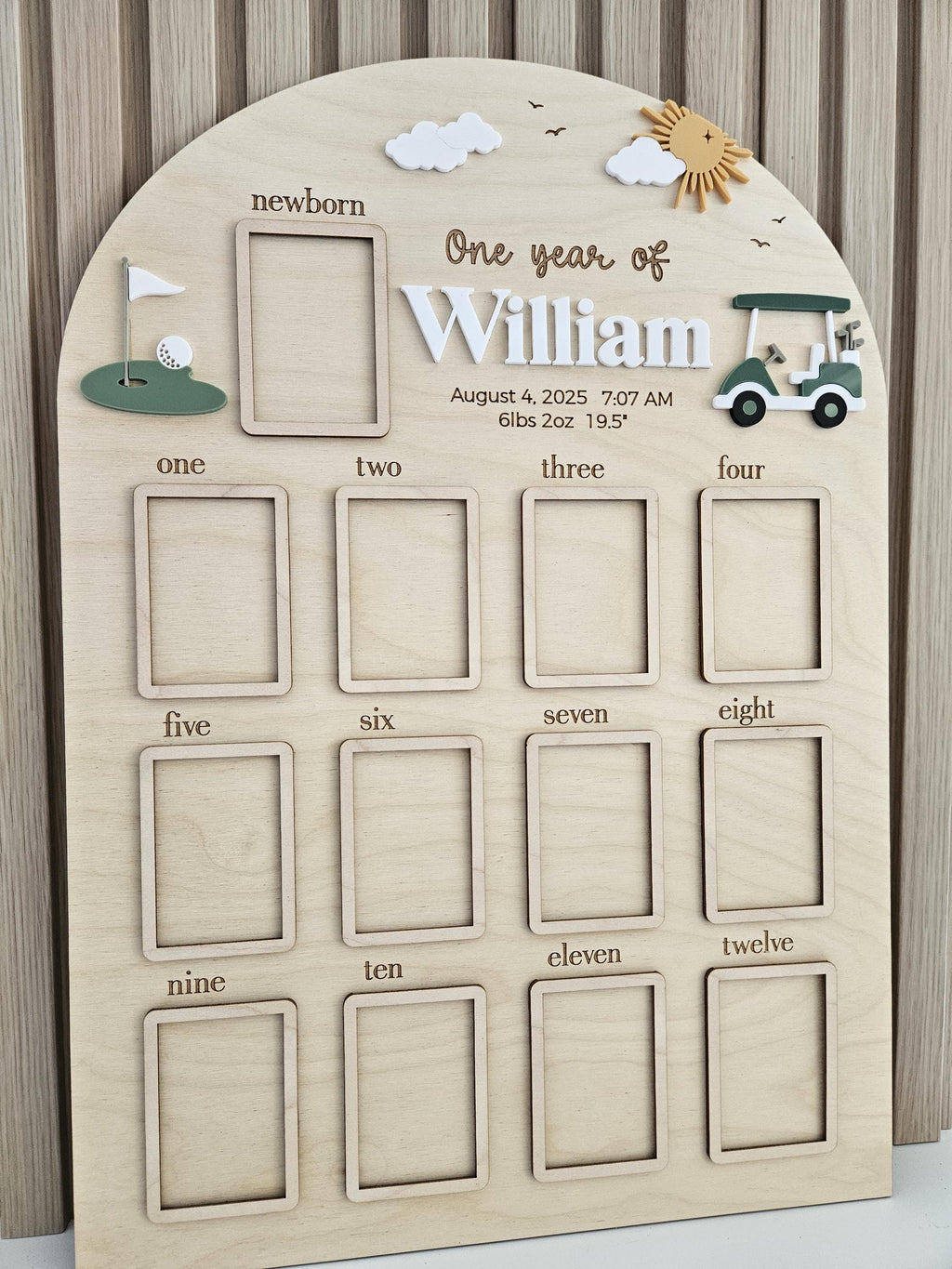 May include: An arched wooden milestone board featuring twelve rectangular photo frames labeled from newborn to twelve. The board includes 3D accents of a golf green, a golf cart, clouds, and a sun, with the text One year of William, August 4, 2025, 7:07 AM, 6lbs 2oz, 19.5 inches.