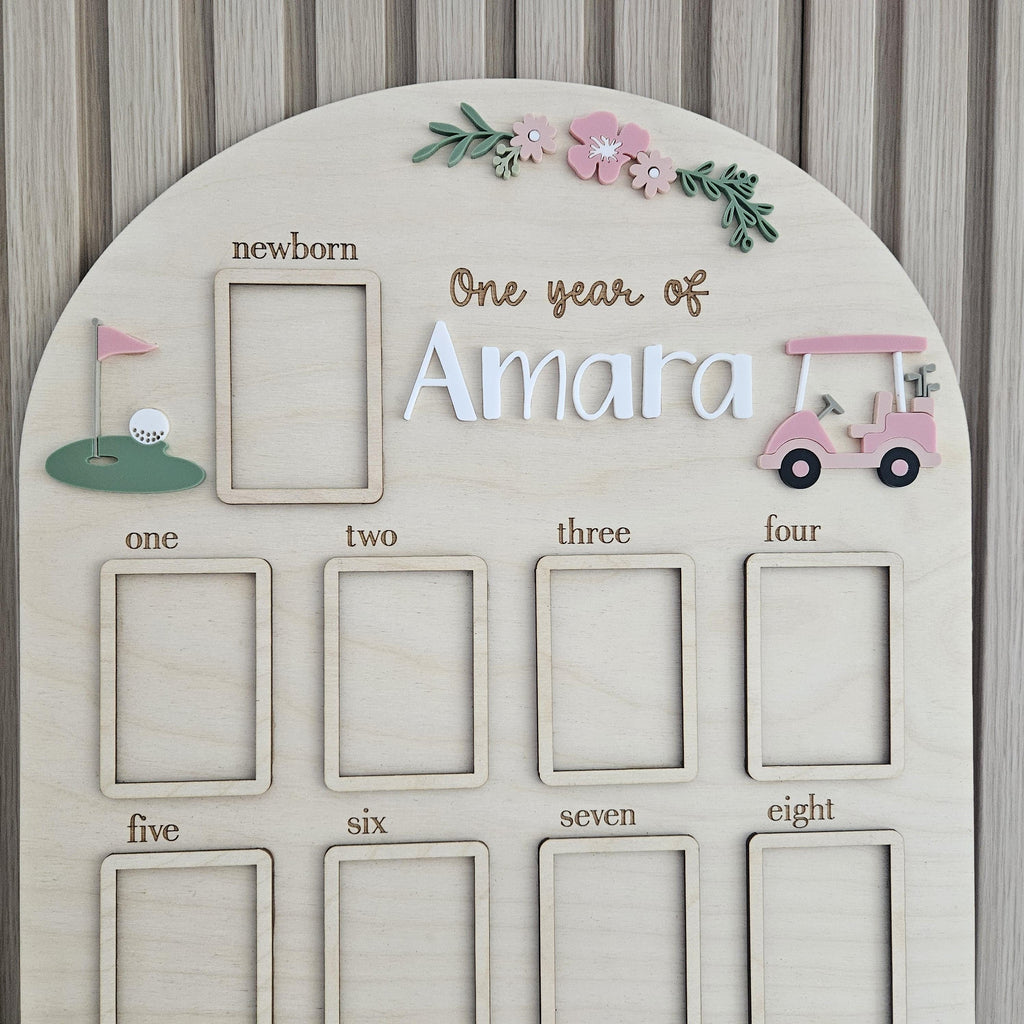 May include: An arched wooden milestone board featuring the text One year of Amara, with twelve rectangular photo frames labeled from newborn to eight. The board is decorated with a pink golf cart, a golf flag, and floral accents in pink and green.