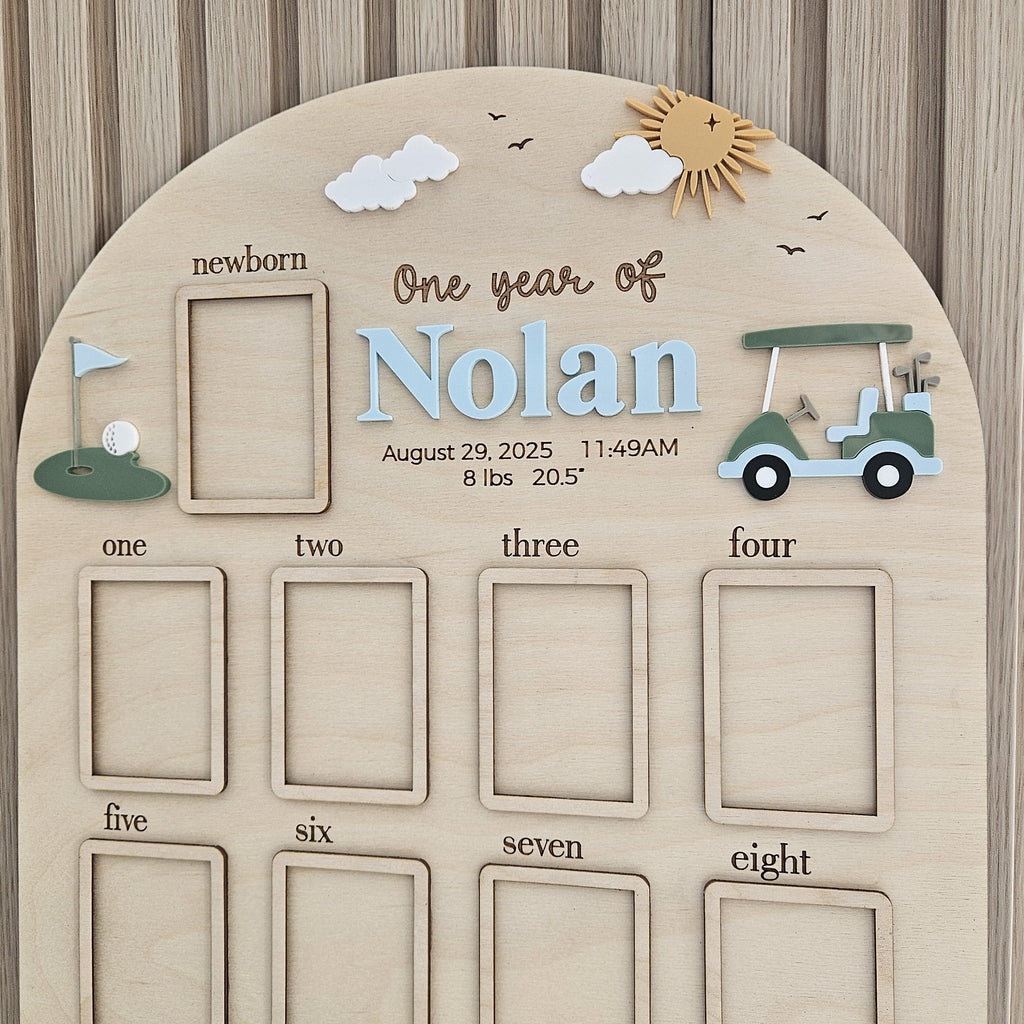 May include: An arched wooden milestone board featuring the text One year of Nolan, August 29, 2025, 11:49AM, 8 lbs, 20.5 inches. It includes twelve rectangular photo frames, a golf cart, a golf hole with a flag, white clouds, and a sun.
