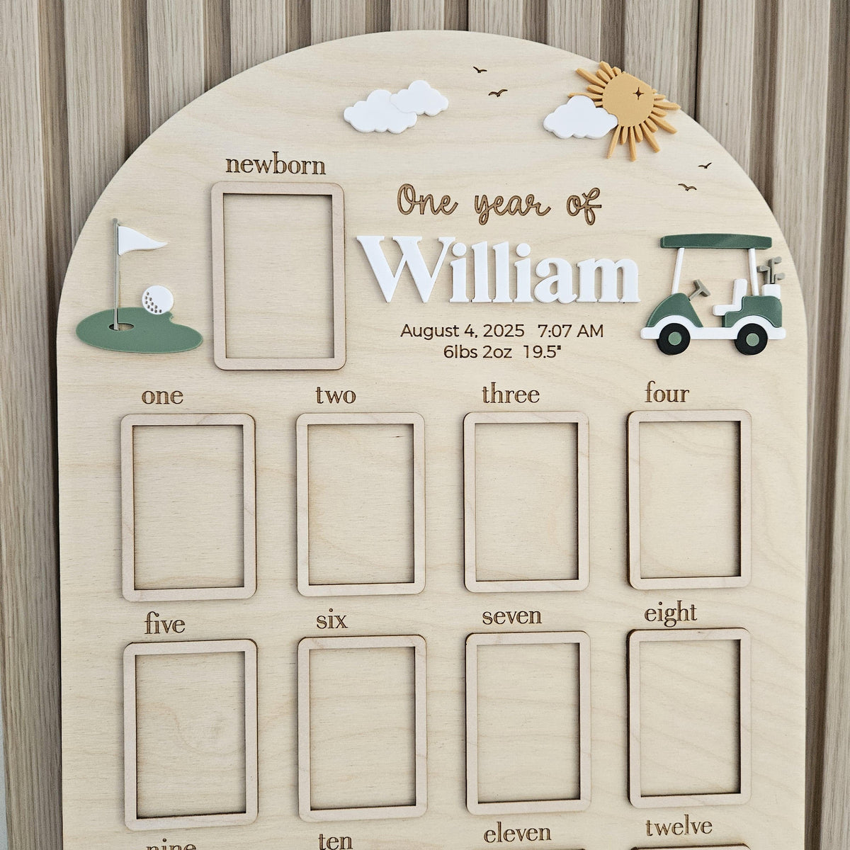 May include: An arched wooden milestone board featuring the text One year of William, August 4, 2025, 7:07 AM, 6lbs 2oz, 19.5 inches. It includes a newborn frame, twelve monthly photo slots, a golf cart, a golf hole, a sun, and white clouds.