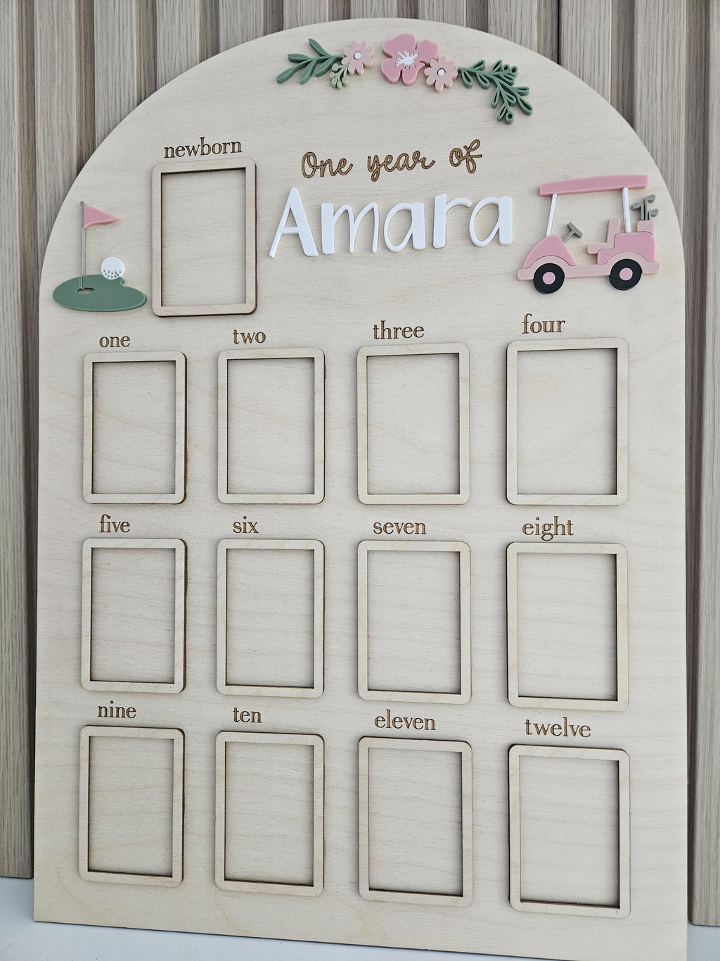May include: An arched wooden milestone board featuring the text One year of Amara. It includes thirteen rectangular photo frames labeled from newborn to twelve. Decorative elements include a pink golf cart, a green putting green, and flowers.