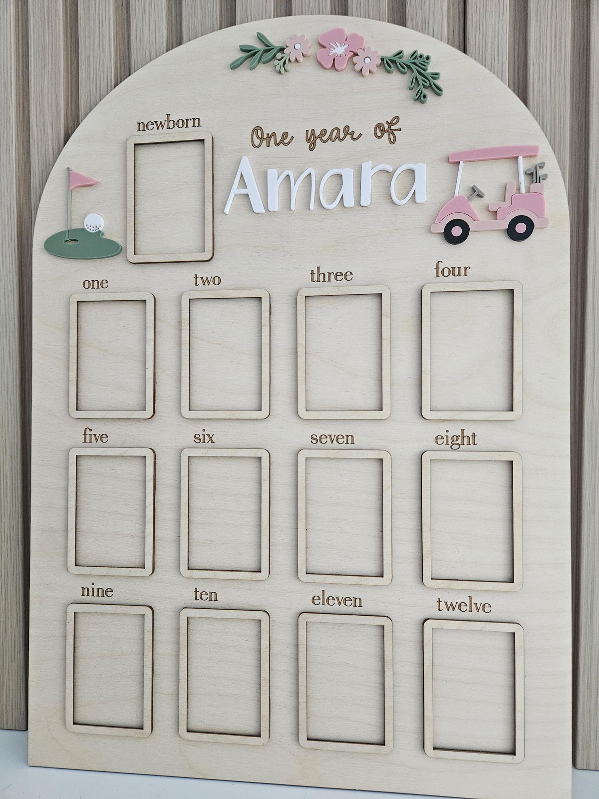 May include: An arched wooden milestone board featuring the text One year of Amara. It includes thirteen rectangular photo frames labeled from newborn to twelve. Decorative elements include a pink golf cart, a green putting green, and flowers.