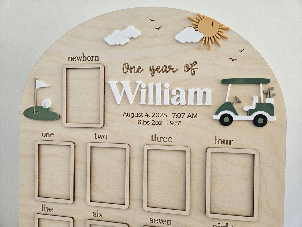 May include: An arched wooden milestone board featuring the text One year of William, August 4, 2025, 7:07 AM, 6lbs 2oz 19.5". The board includes twelve rectangular photo frames, a golf cart, a golf hole with a flag, clouds, and a sun.