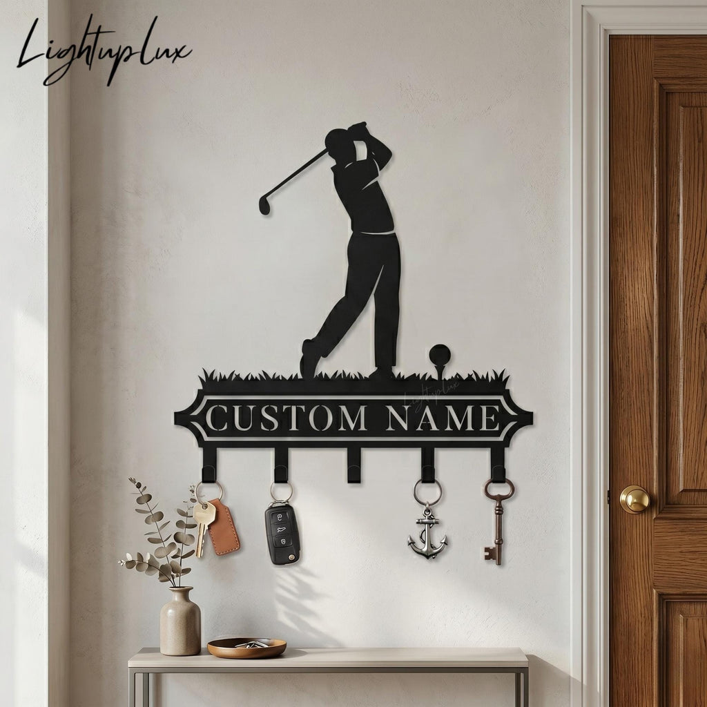 May include: Black metal key holder featuring a golfer silhouette in mid-swing. The key holder has hooks below a banner that reads "CUSTOM NAME". Keys and keychains hang from the hooks. The wall art is mounted on a light-colored wall.