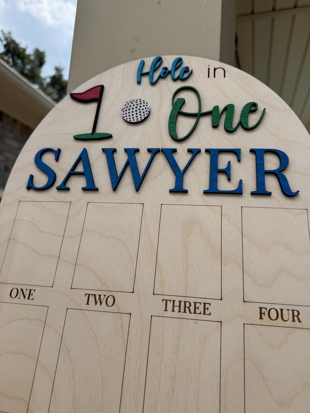 May include: Wooden golf-themed sign with the words "Hole in One" and the name "Sawyer" in blue and green lettering. Includes a golf ball and flag graphic. Features spaces labeled "One," "Two," "Three," and "Four."